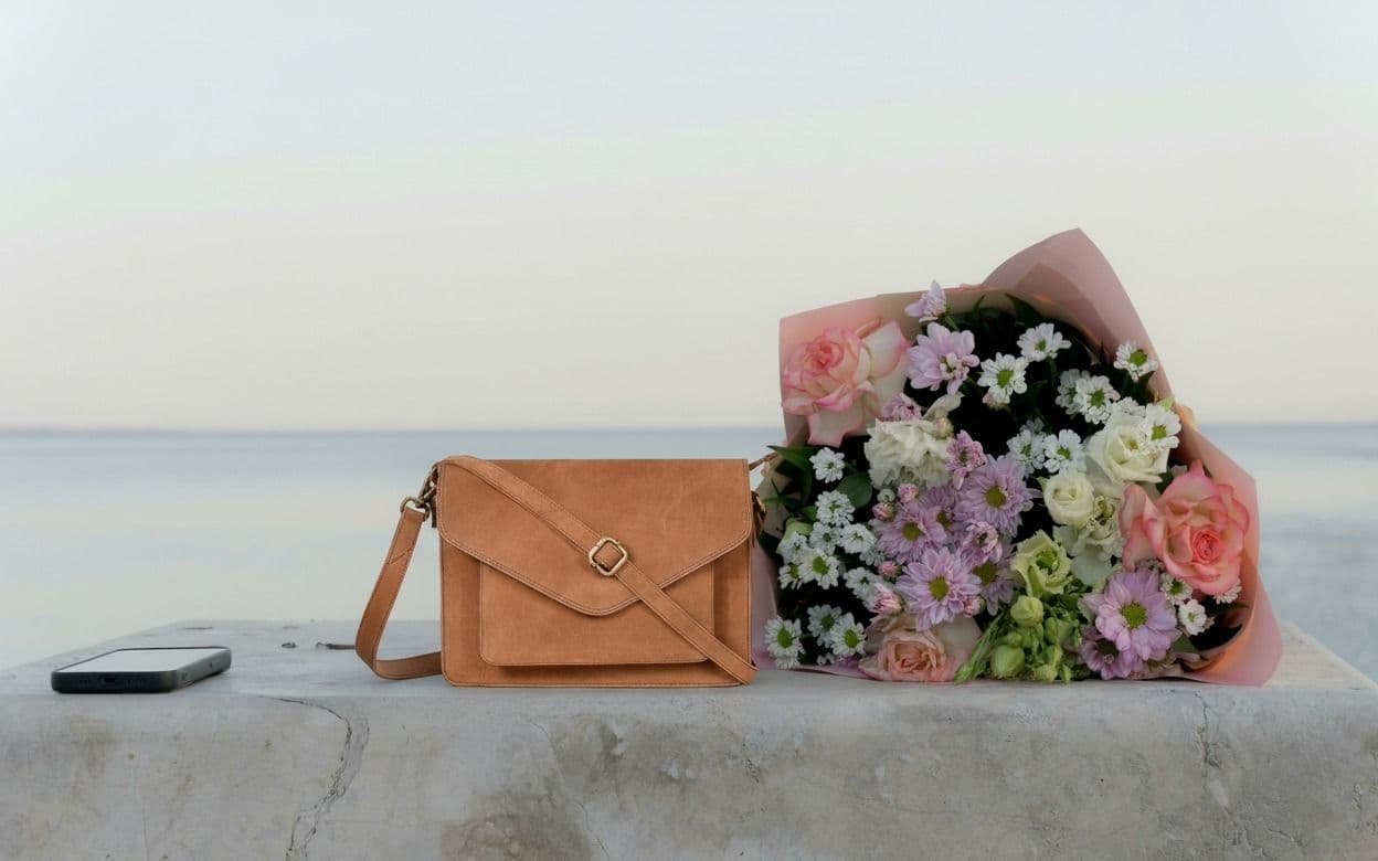Tan flap crossbody bag with gold buckle displayed beside a bouquet of flowers on a stone surface by the sea.